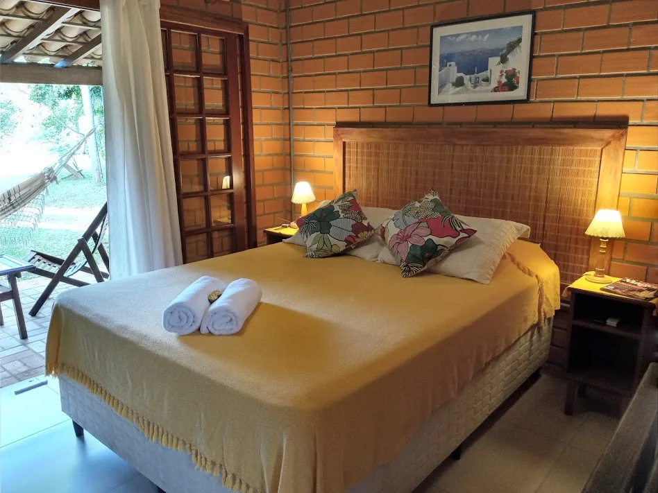 Suíte com cama de casal, parede de tijolinho e porta aberta para a varanda com rede