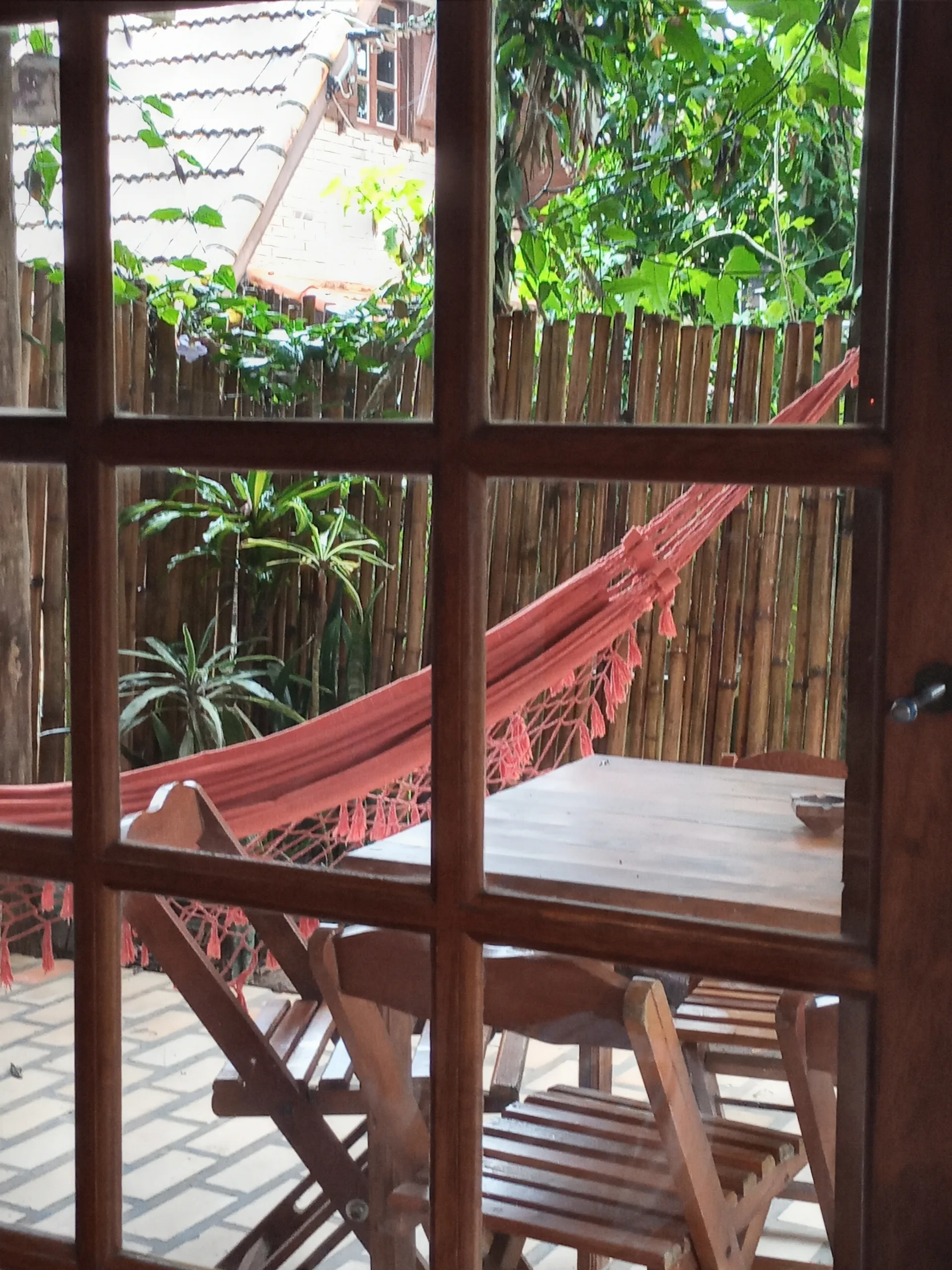 Vista pela janela da cozinha dos hóspedes para a varanda com rede, mesa e cadeiras de madeira