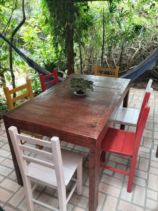 Mesa com cadeiras coloridas e rede na varanda, de frente para a mata